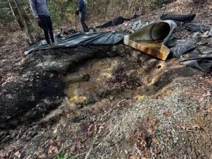 Workers repairing a stormwater energy dissipater, with a culvert outlet and black tarp covering the ground, rocks scattered around, and a bucket nearby in a forested area.