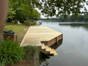 A newly constructed wooden dock in Raleigh, NC, stretches over a tranquil lake, featuring a sturdy platform and steps, framed by greenery and a distant boat.