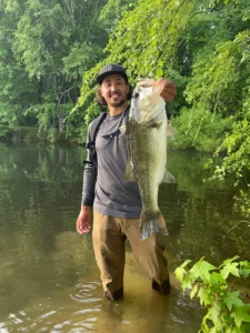 A proud angler stands in shallow water surrounded by lush greenery in Raleigh, holding a large fish, showcasing the success of local fish stocking efforts, with trees and reflections visible in the calm lake.
