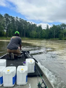 A person in a green cap works on a boat, administering a Calcis Treatments, while moving through a pond with murky water. Green trees line the horizon under a cloudy sky, with fishing gear visible on the boat.