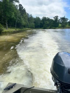 A boat with a motor cuts through a pond, creating a wake with foamy water. The pond water appears cloudy on one side, surrounded by lush green trees under a partly cloudy sky.