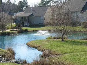 Residential stormwater pond with fountain, grassy shoreline and surrounding homes.